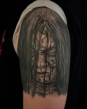 Black and grey portrait of a long-haired horror figure on an upper arm — by Adam Crane